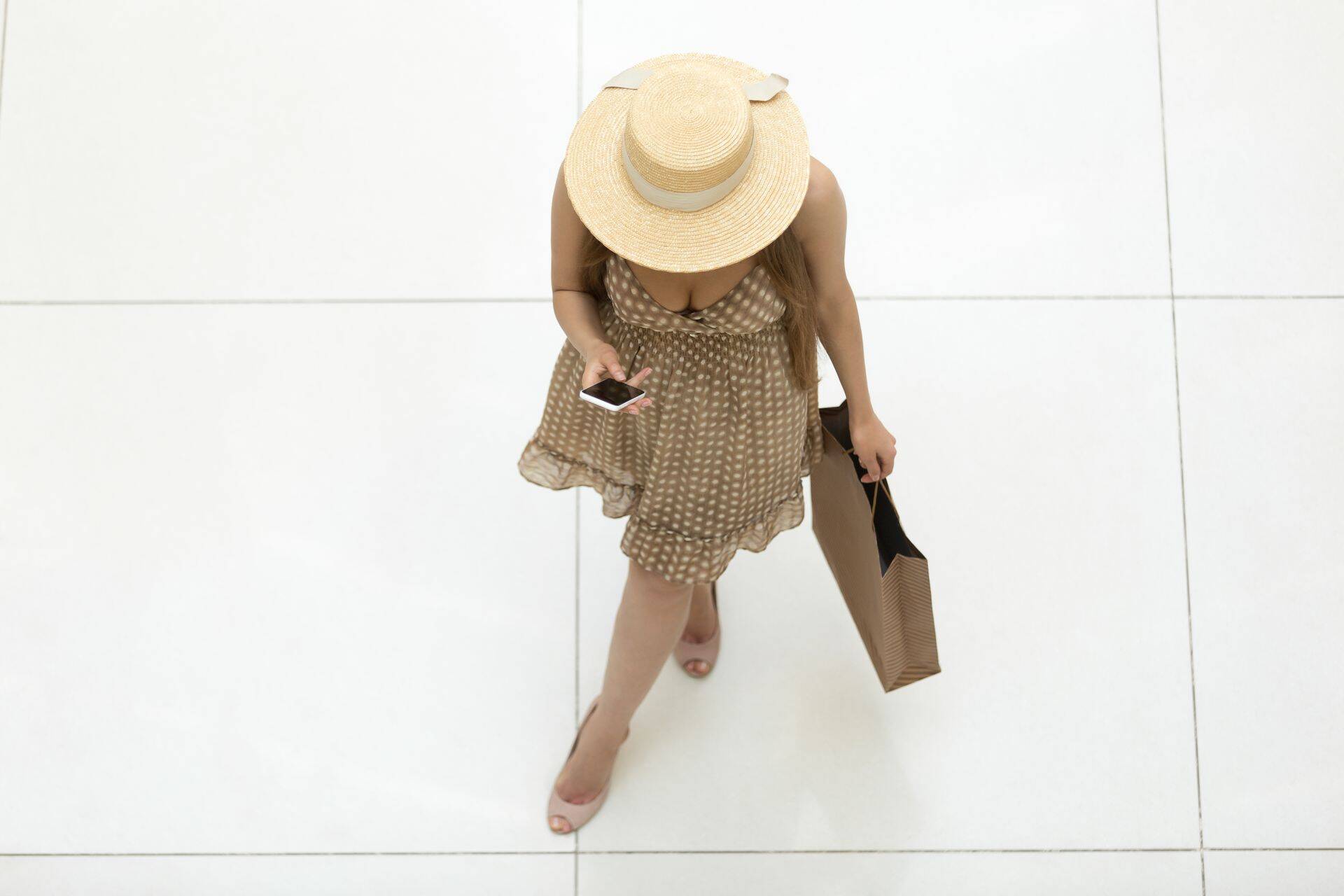 Young pretty shopper woman wearing hat and polka dot dress standing in shopping centre carrying shopping bag and holding smartphone, touching screen, reading message. View from above