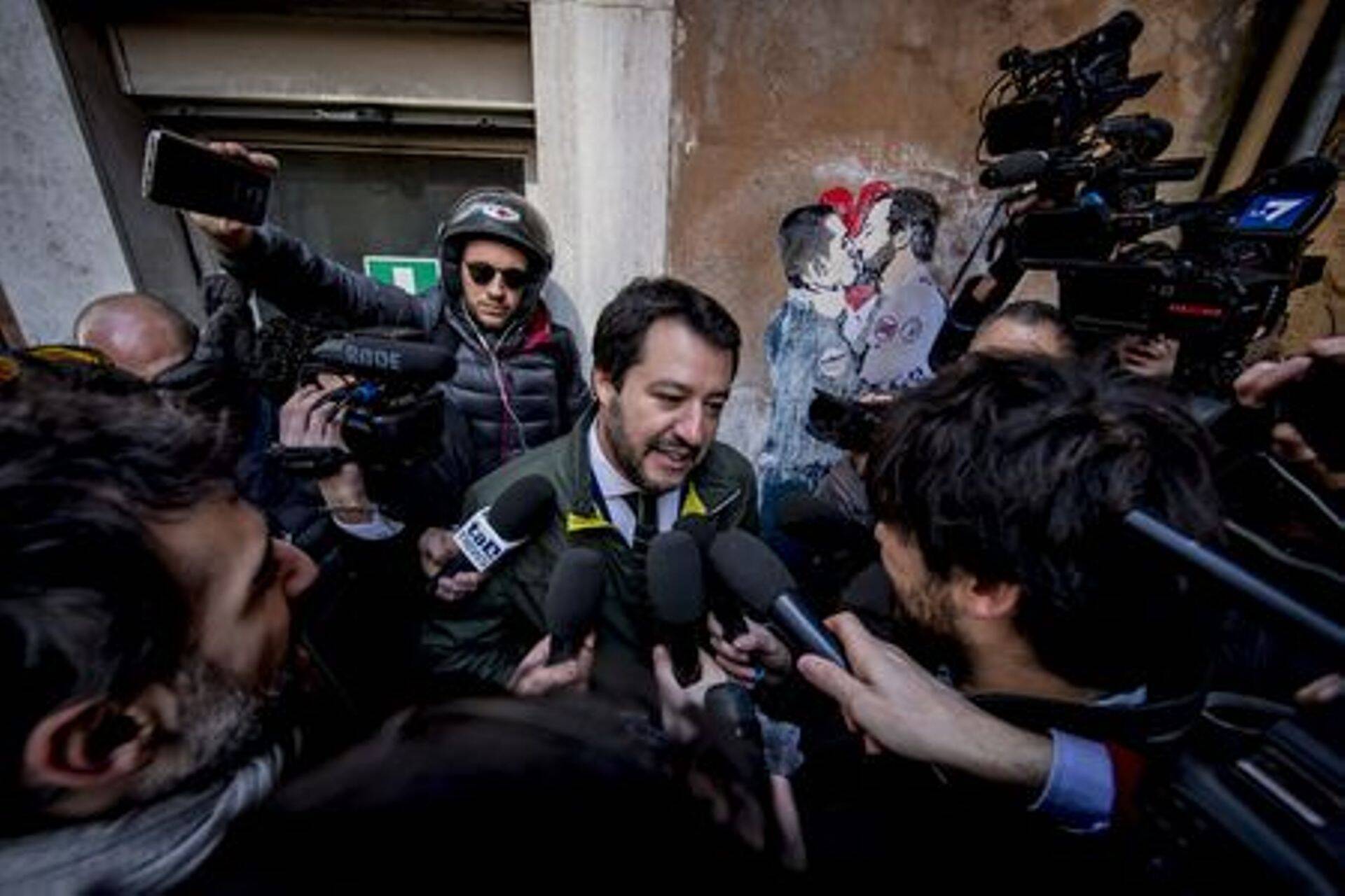 Liiga juht Matteo Salvini ajakirjanikele vastuseid andmas, taustal grafiiti, mis kujutab teda ja M5S juhti üksteise embuses.