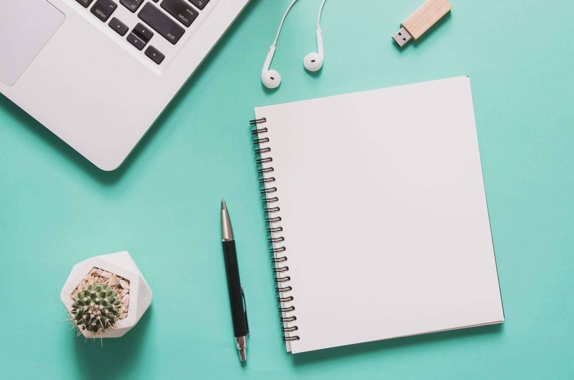 Office workplace concept. Computer laptop with blank notebook, cactus, pen, flash drive, earphone on blue background.