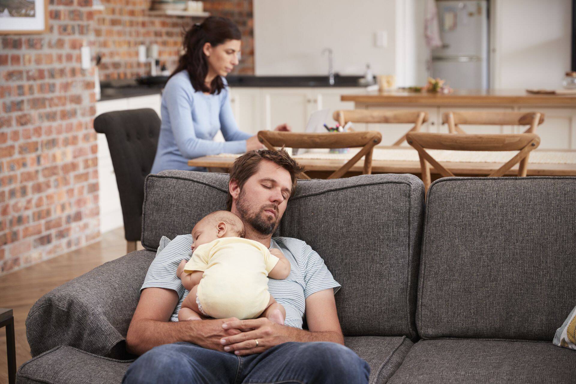 Tired Father With Baby Son Asleep On Sofa As Mother Works